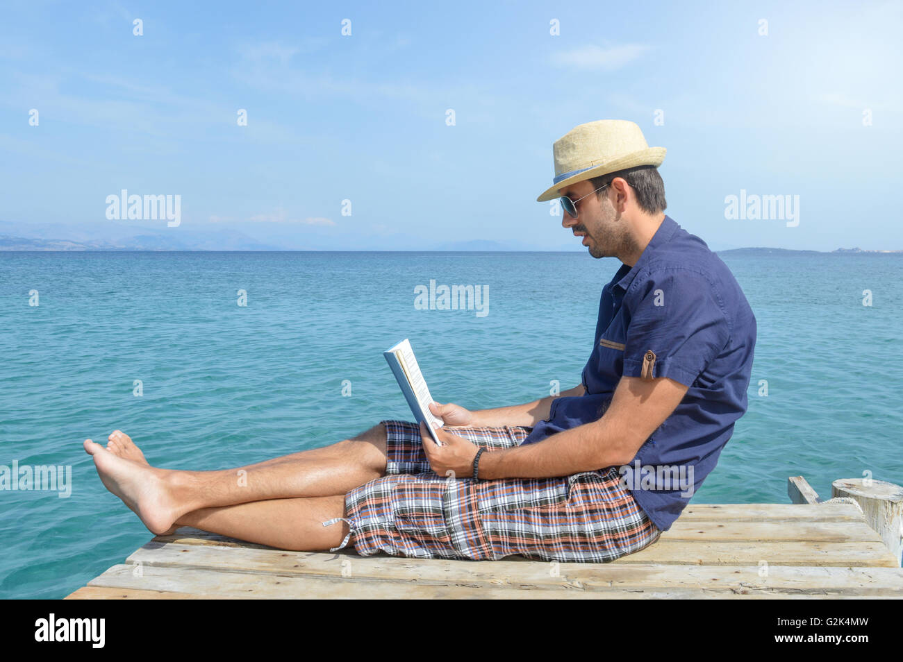 Man in blue shirt sitting on the dock reading a book Stock Photo