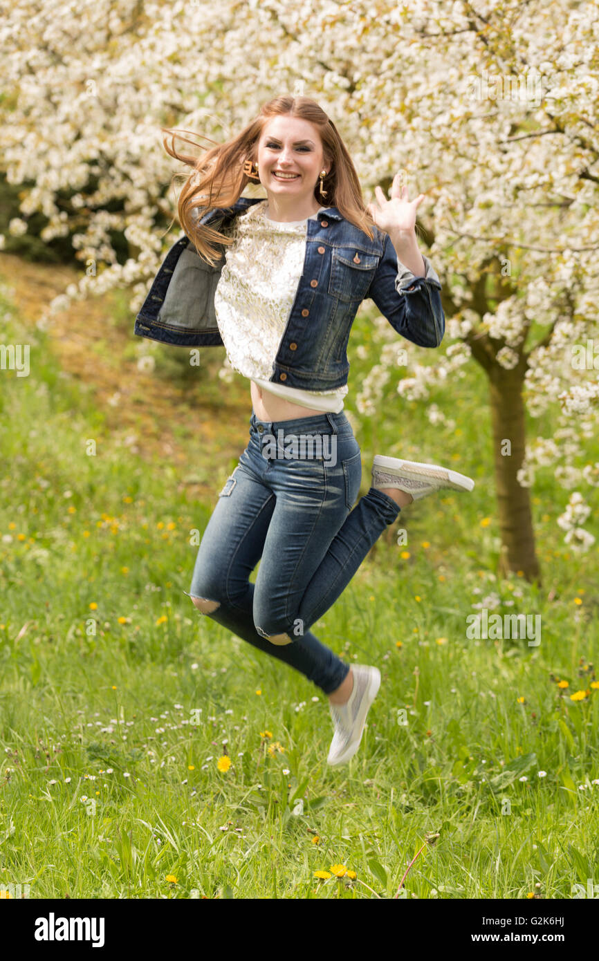 A young brown haired woman in jeans enjoying the first days of spring in a green meadow surrounded by blooming cherry trees Stock Photo