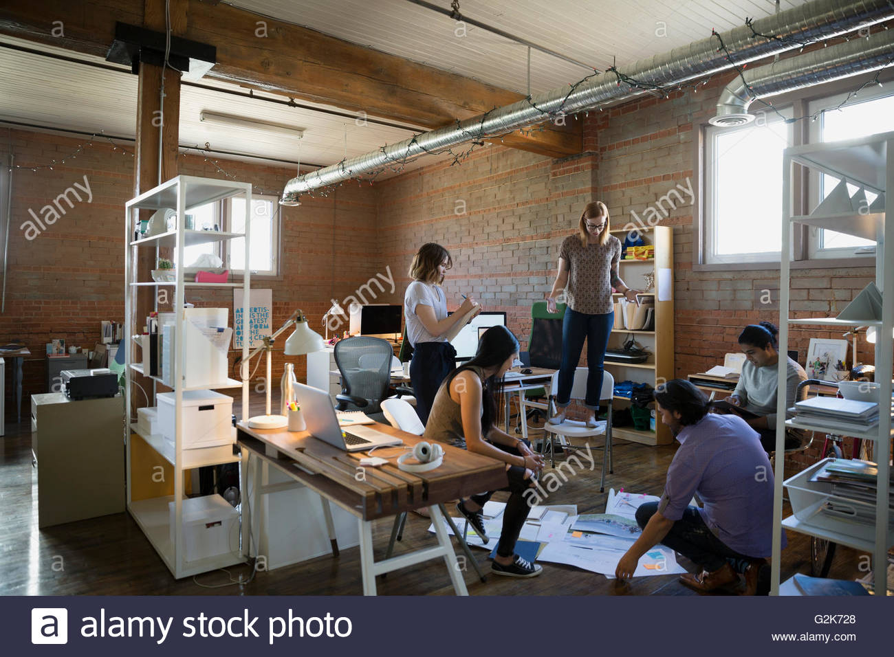 Designer Standing On Chair Looking Down At Proofs And Paperwork On