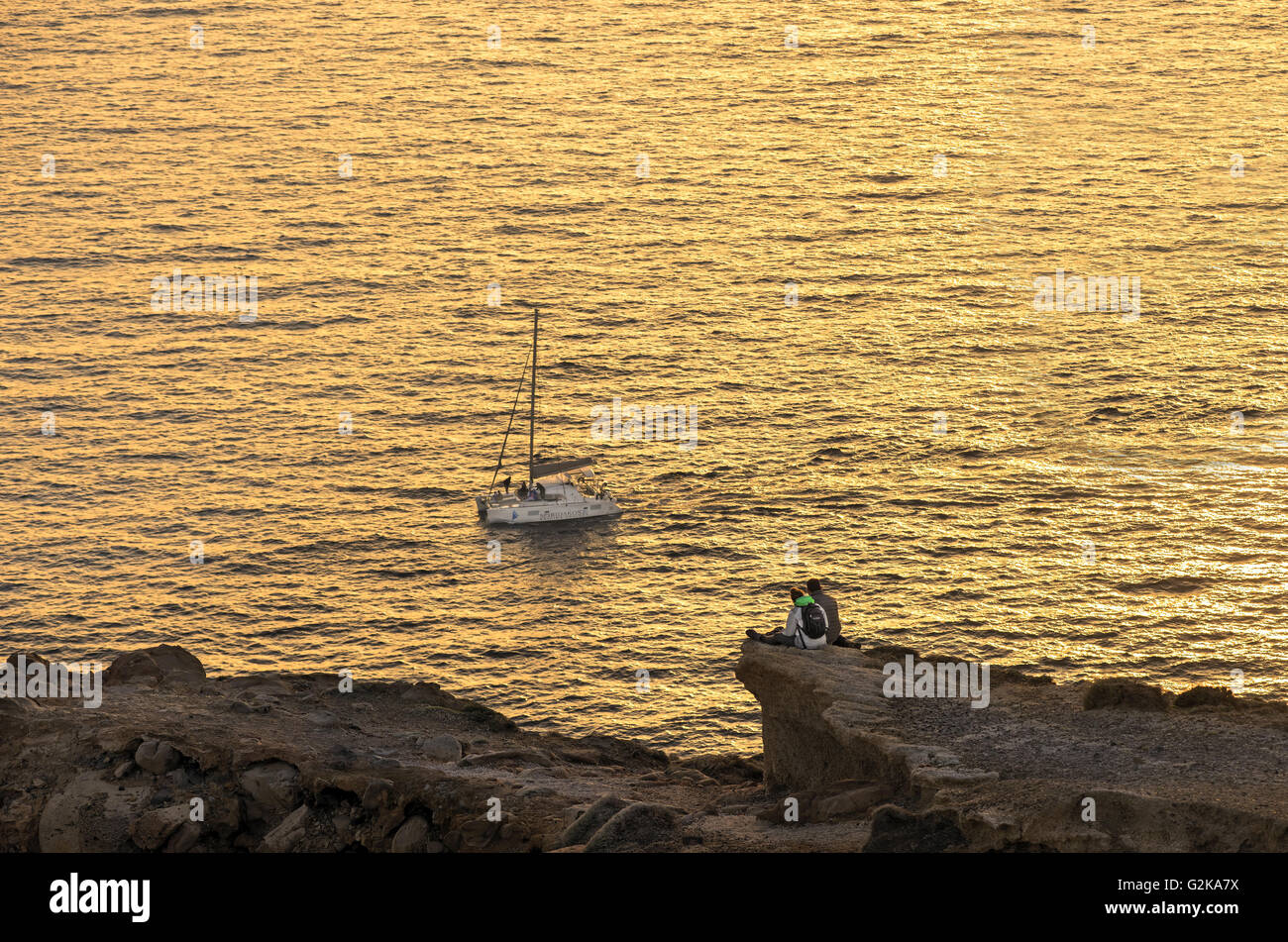 A couple enjoying the sunset sitting at the rocks of Akrotiri with a sailing boat passing in Santorini island, Cyclades, Greece Stock Photo