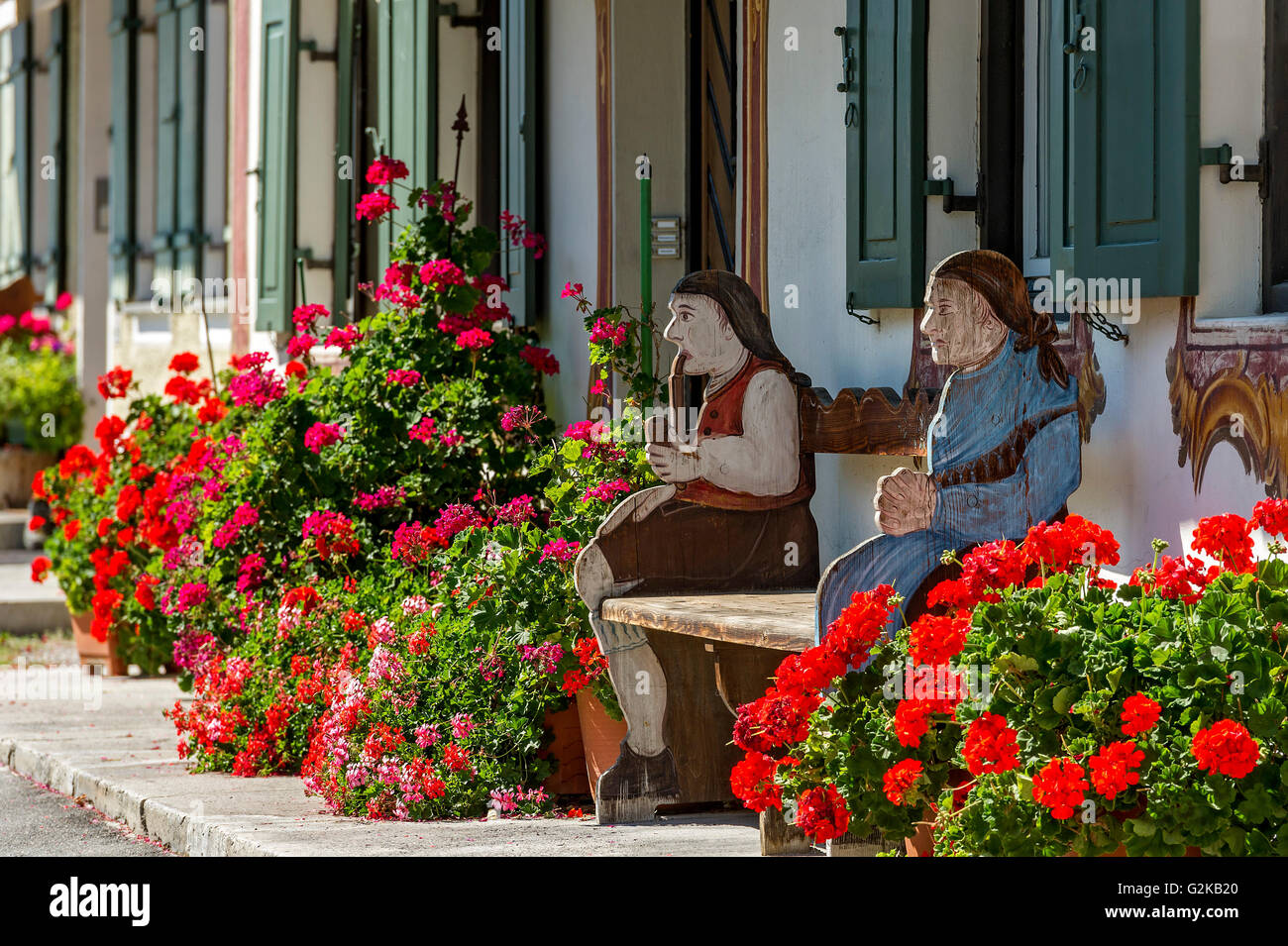 Geraniums (Pelargonium) and traditional Bavarian Bench with figures of farmer and his wife in front of farmhouse, Sun Street Stock Photo
