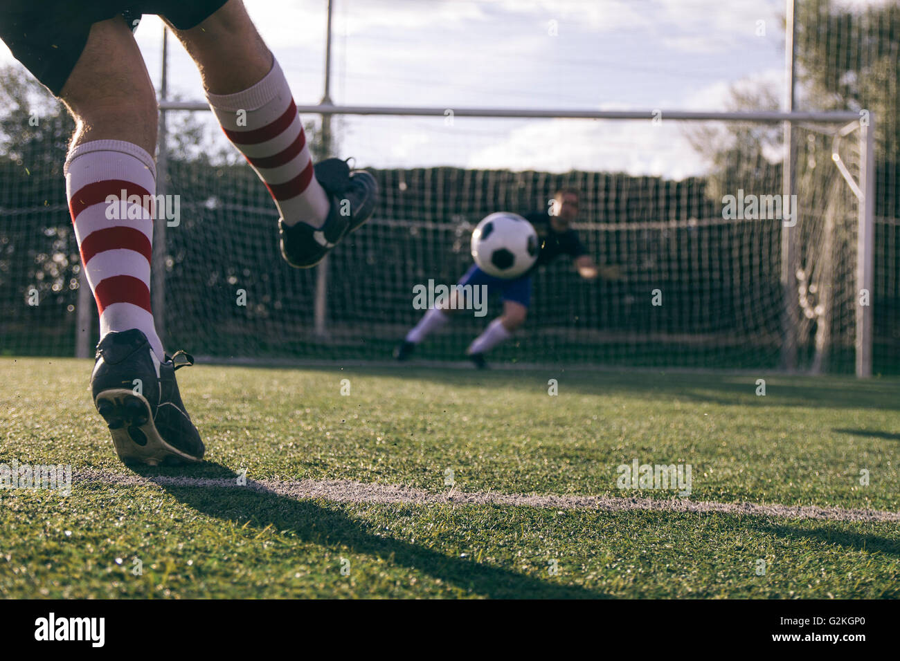 Legs of a footnball player kicking a ball in front of a goal with a goalkeeper Stock Photo