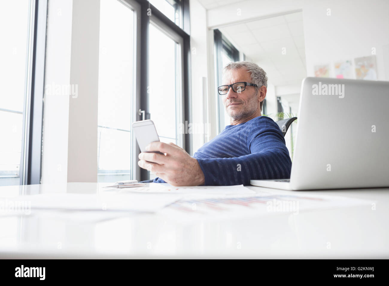 Mature man sitting in office with laptop, using smart phone Stock Photo