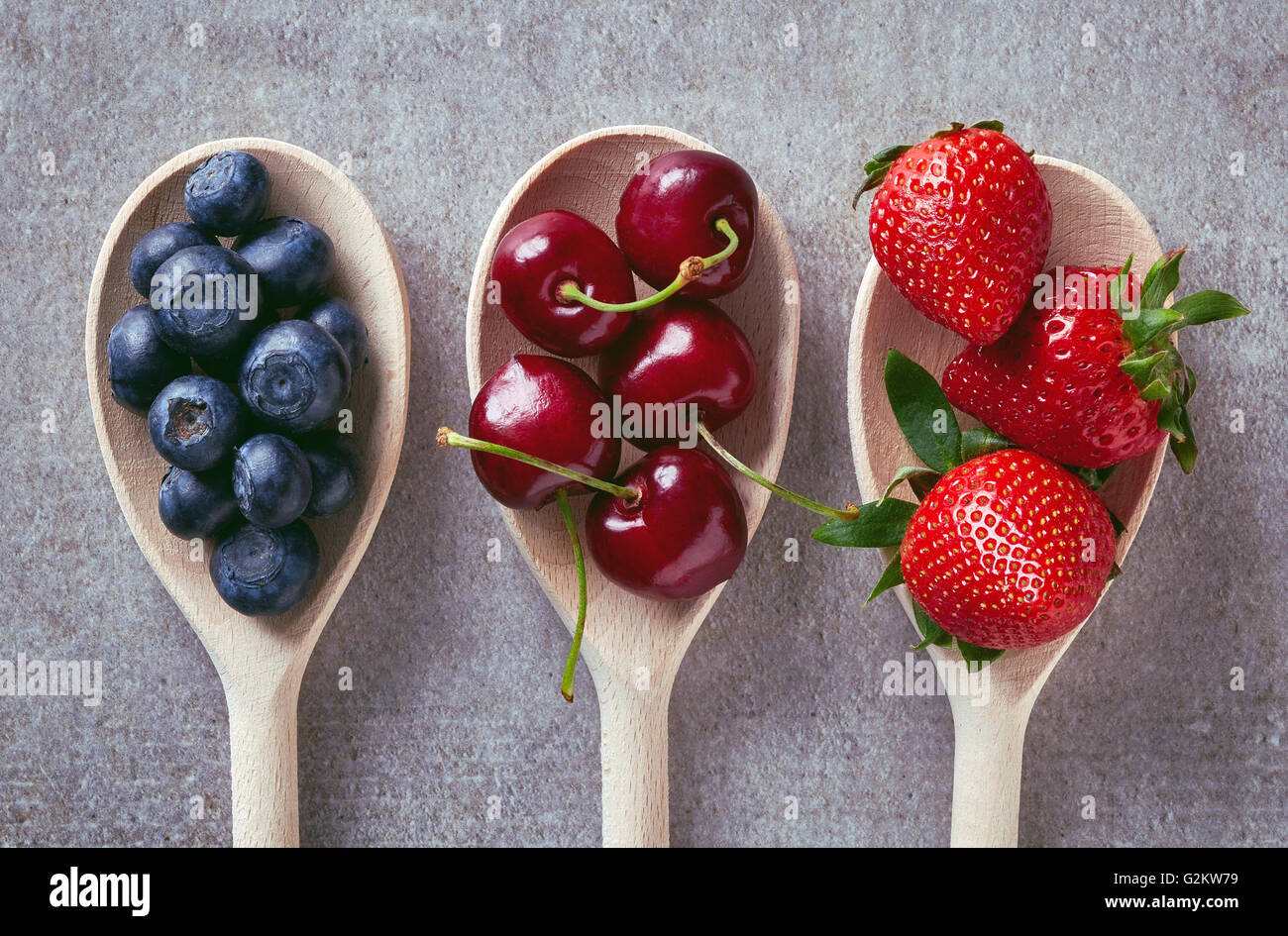 Blueberries, cherries and strawberries in wooden spoons on stone table, top view Stock Photo