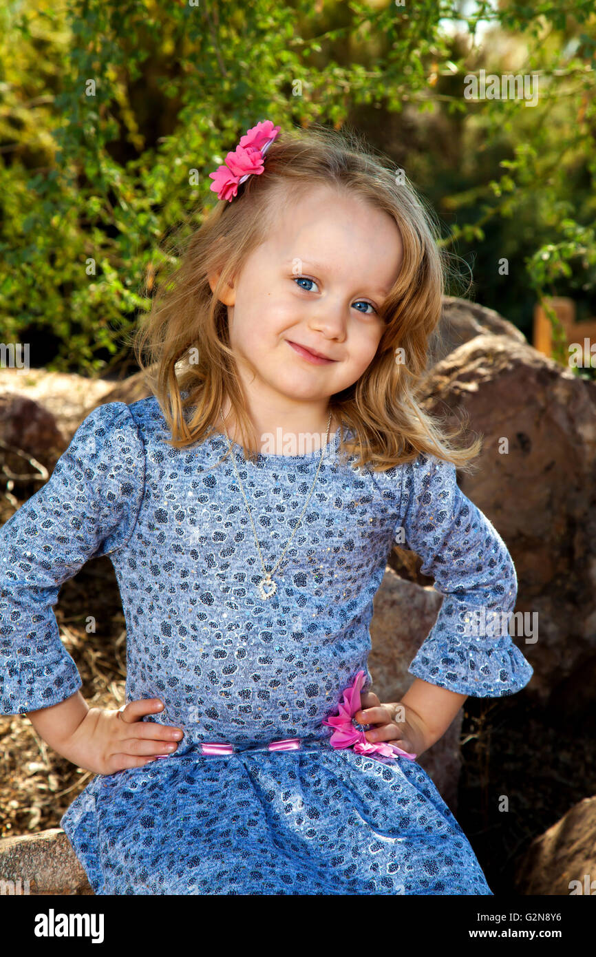 Portrait of a really cute little blond girl with big blue eyes and hands on her hips.  She is wearing blue with pink embellishment Stock Photo