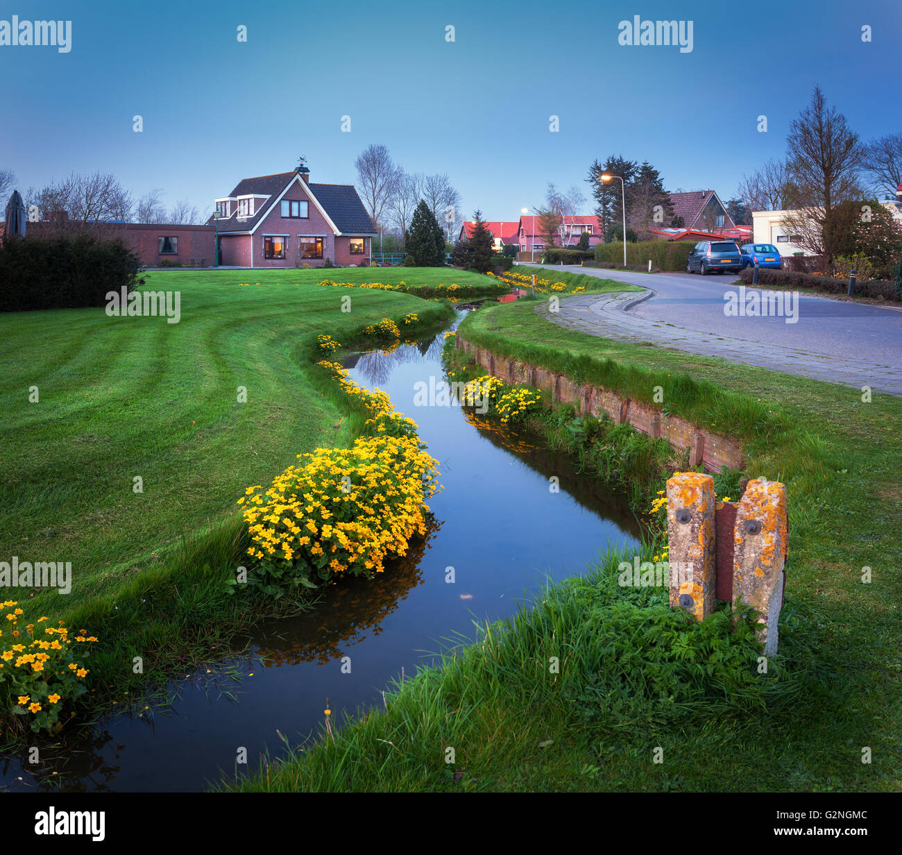 Landscape in dutch village with beautiful house reflected in water canal, courtyard with green grass , yellow flowers and road Stock Photo