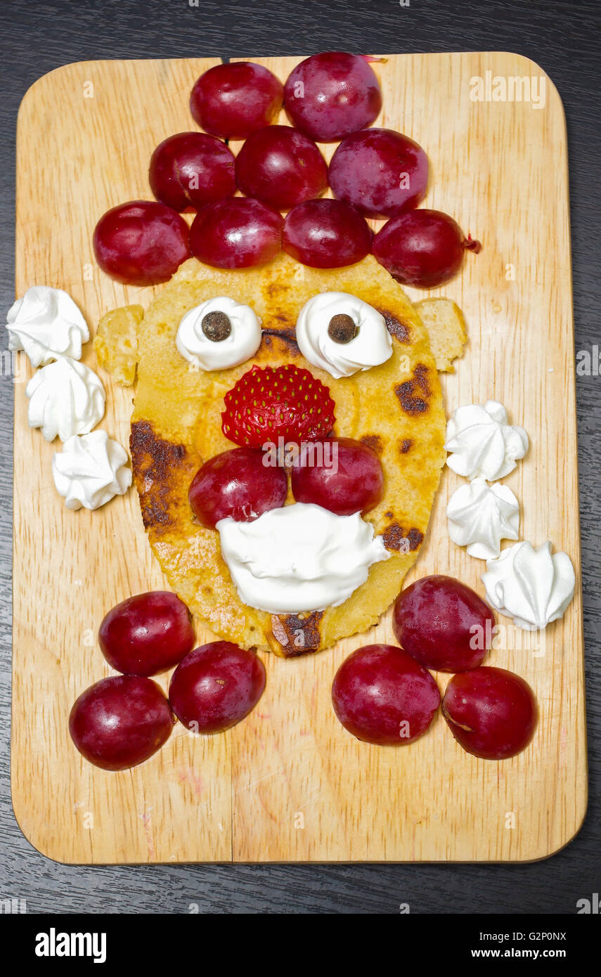 Funny looking face made of sliced grapes, strawberries and pancake, as seen from above Stock Photo