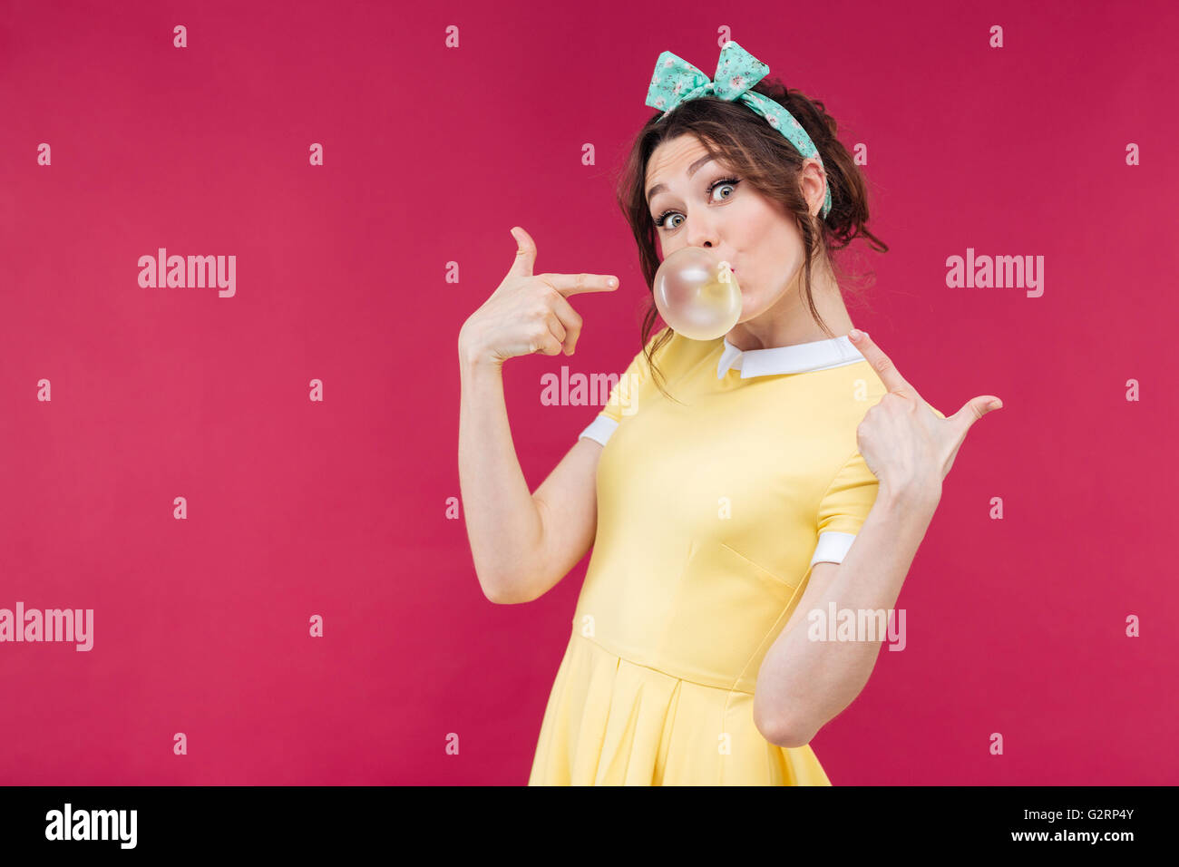 Playful charming young woman in yellow dress pointing on bubble gum balloon over pink background Stock Photo