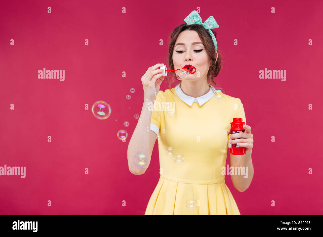 Cute lovely young woman in yellow dress blowing soap bubbles over pink background Stock Photo