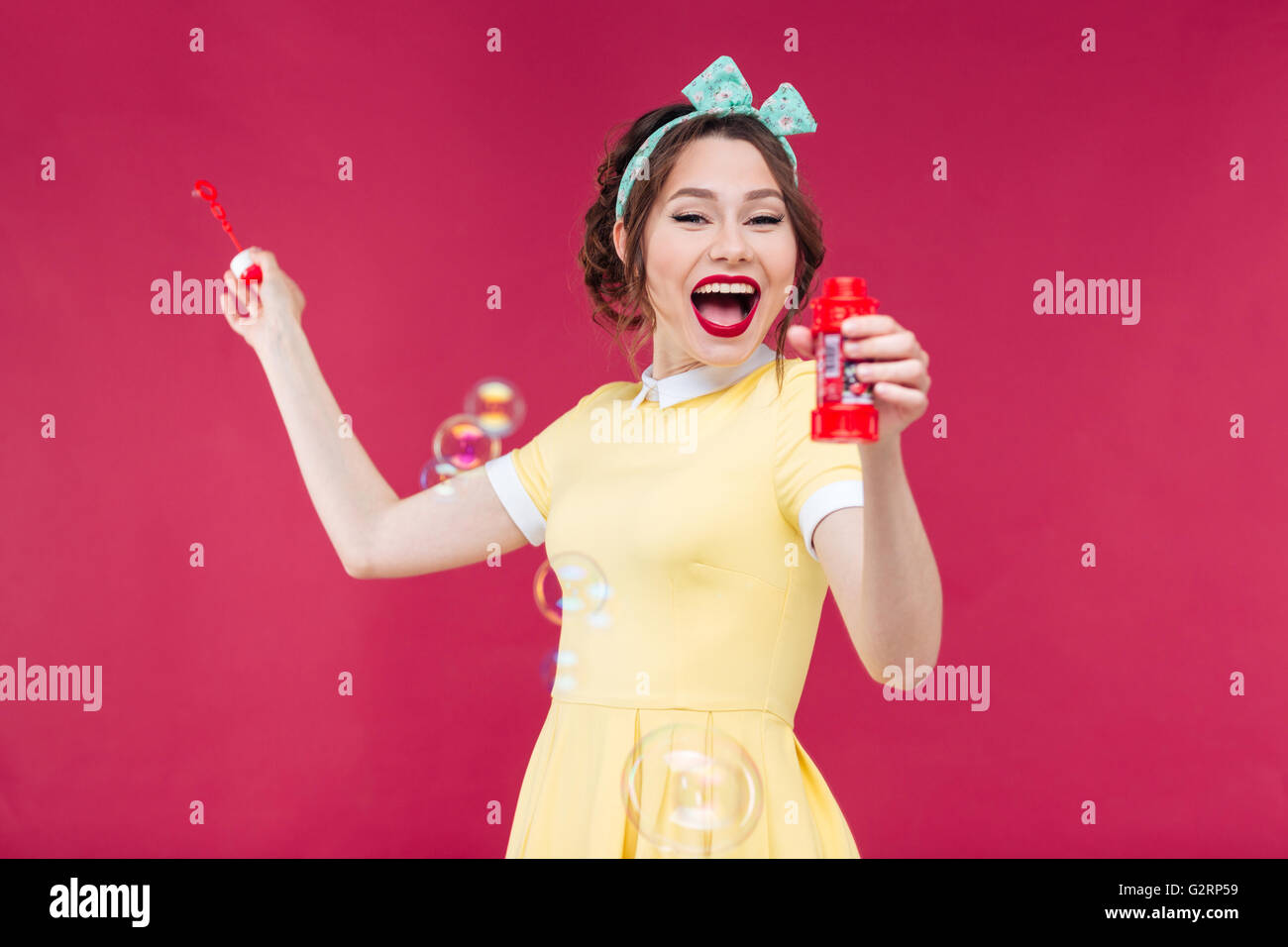 Happy excited pinup girl in yellow dress blowing soap bubbles and laughing over pink background Stock Photo