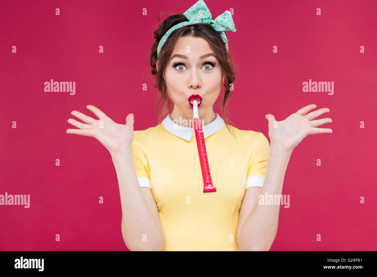 Funny lovely pinup girl blowing in party whistle and holding copyspace on palms over pink background Stock Photo