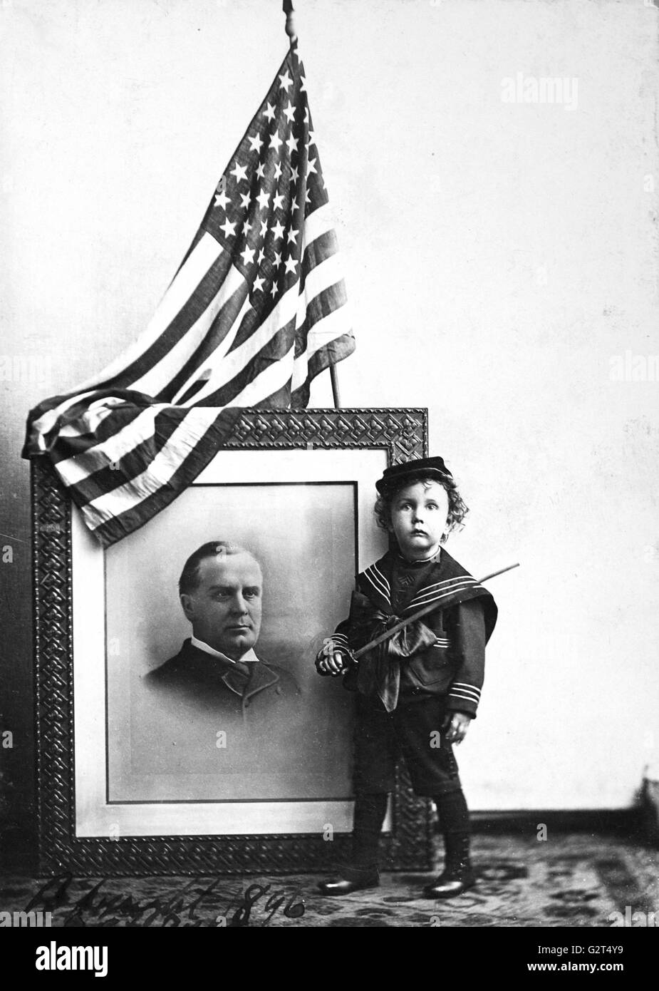 Patriotic, sentimental photo of a child dressed in a military outfit posed with a portrait of Pres. William McKinley and an American flag. Stock Photo
