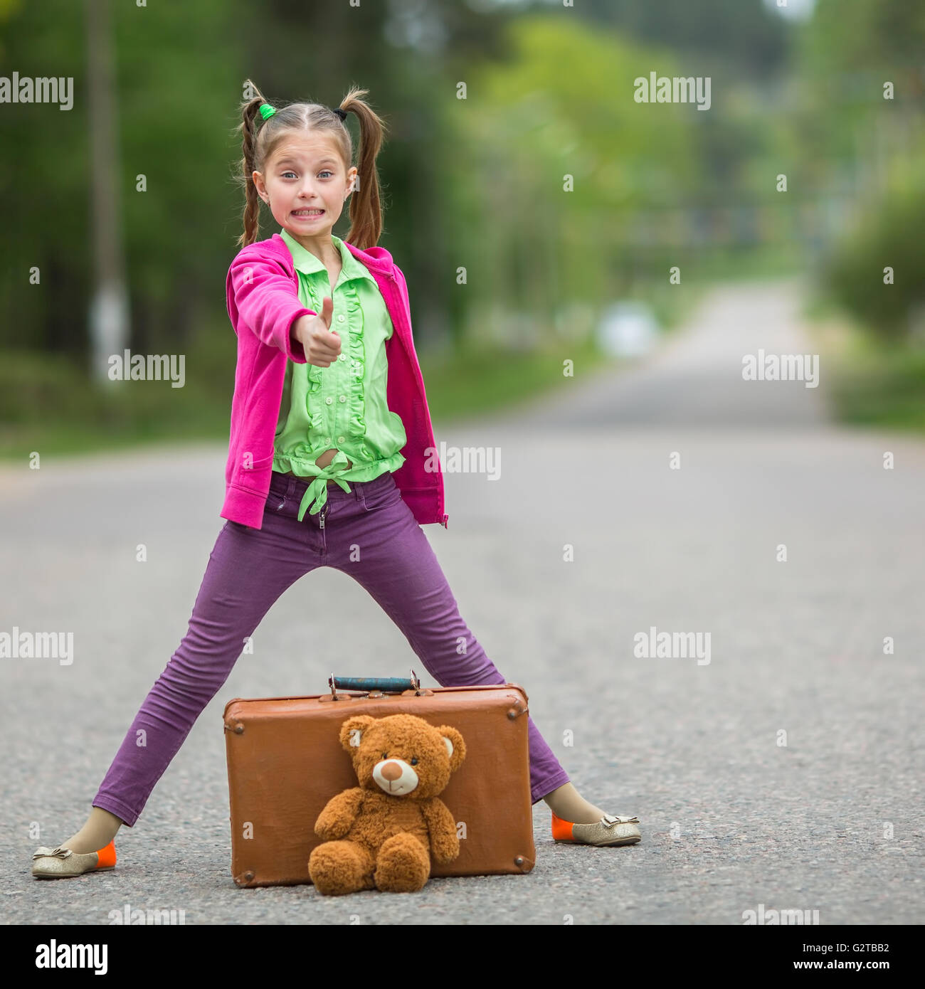 Little girl in bright clothes standing on the road with a suitcase and ...