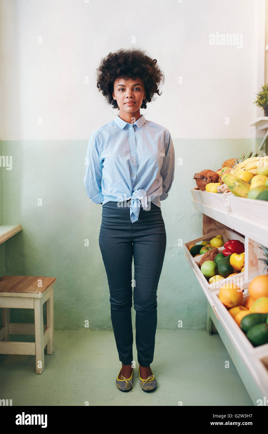 Full length portrait of young african woman standing in a juice bar. Juice bar owner standing by a shelf with fruits. Stock Photo