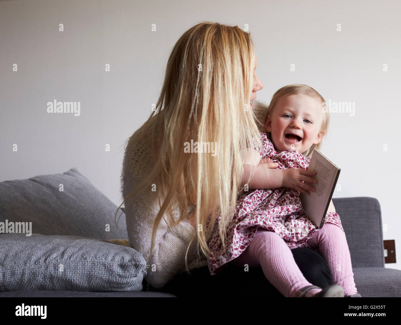 Mother Sitting On Sofa Reading Story With Daughter Stock Photo