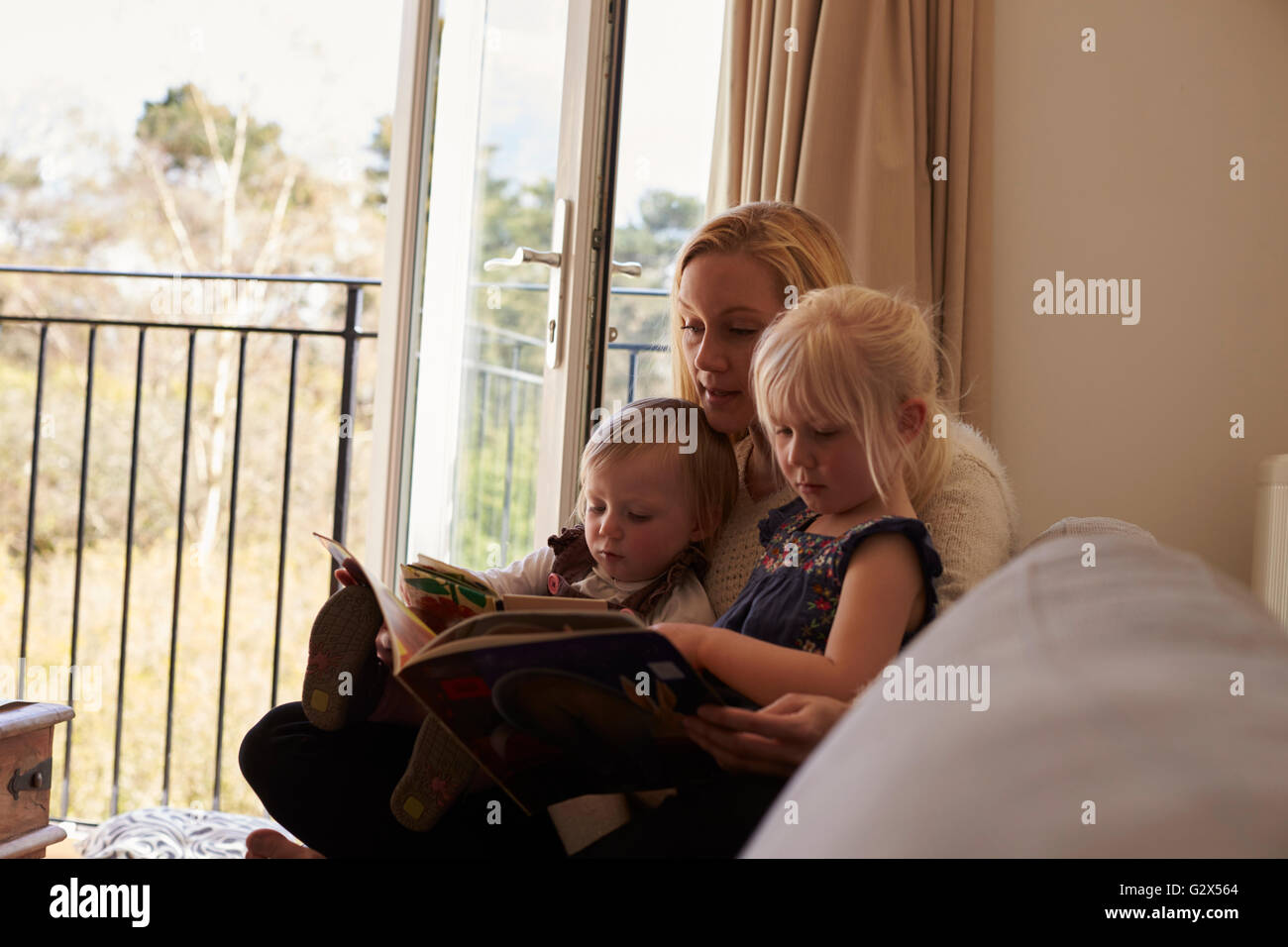 Mother Sitting On Sofa Reading Story With Daughters Stock Photo