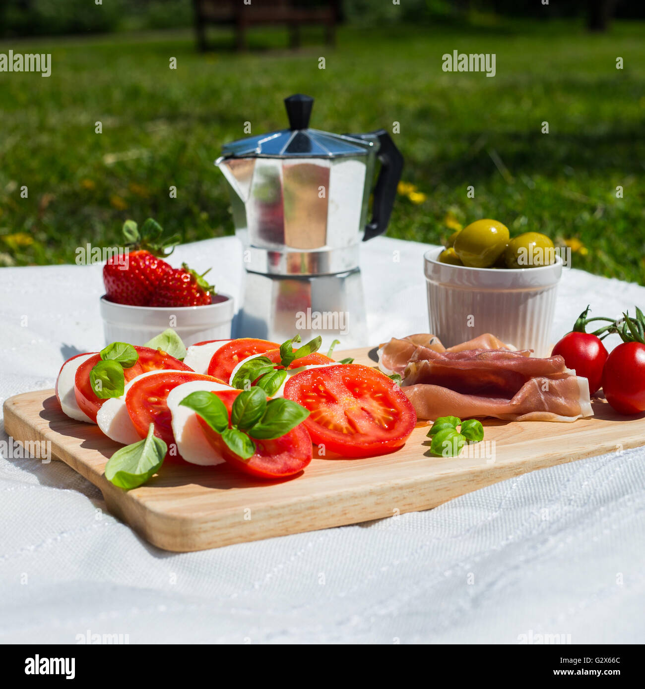 A picnic of tomato and mozzarella salad, parma ham, strawberries, olives and espresso pot in a park Stock Photo