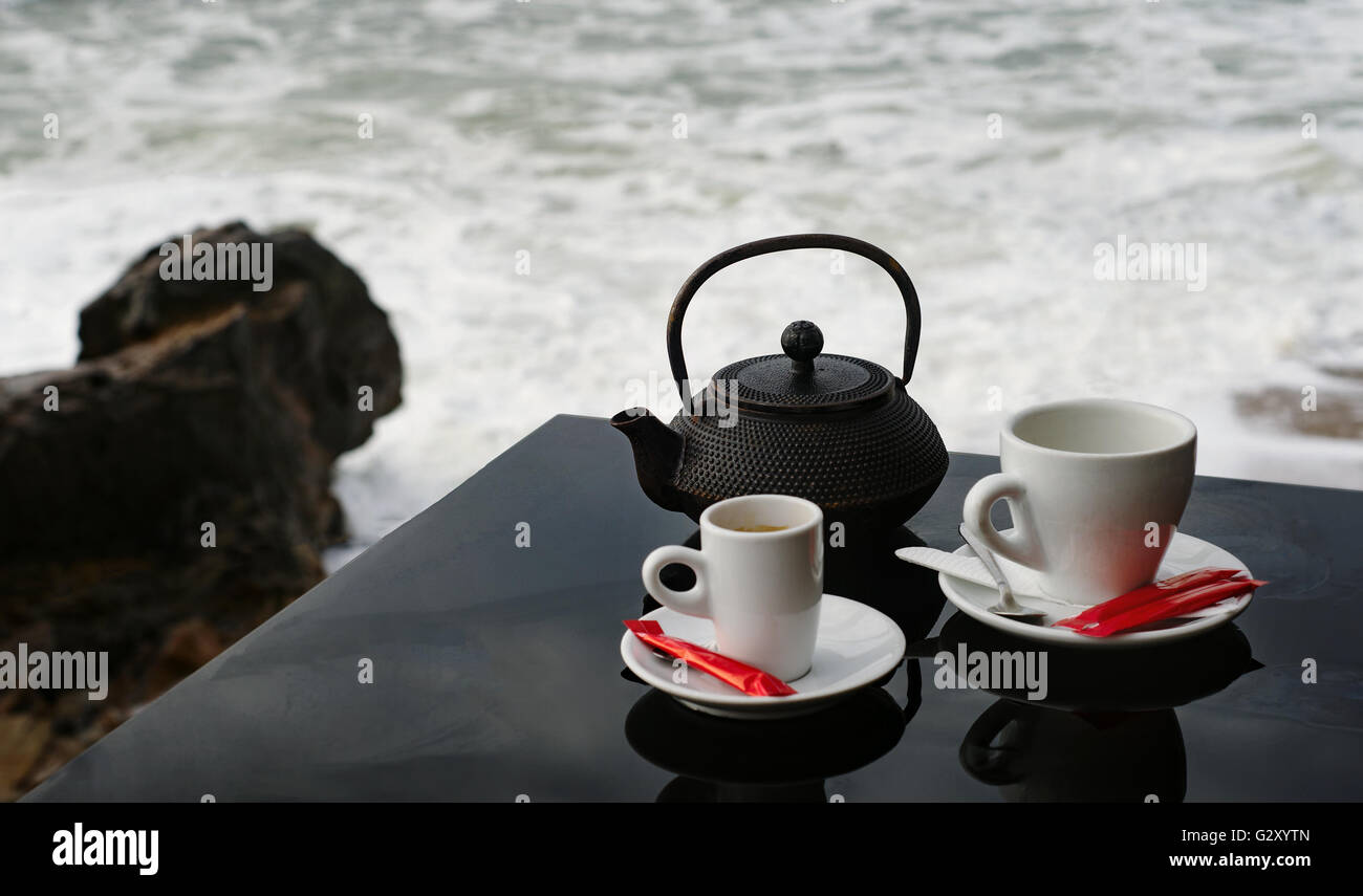 Tea and coffee cup pot set service on black shiny table with view of ocean shore Stock Photo