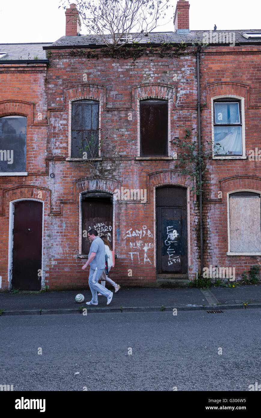 Two people walk along kicking a football in front of a derelict house boarded up with steel plates, written over with graffiti. Stock Photo