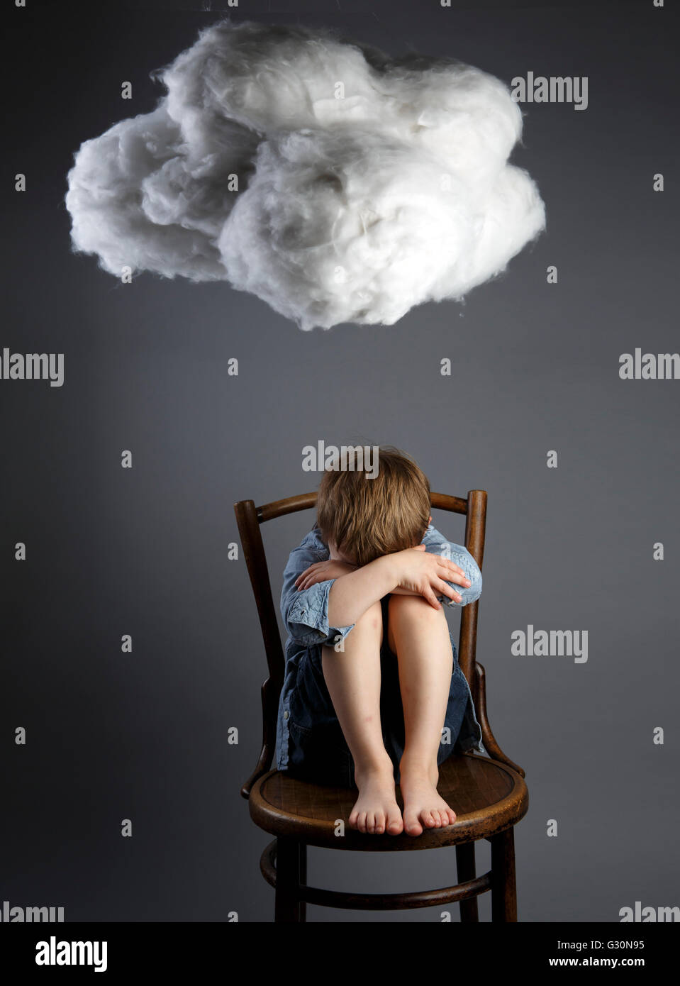 Young boy child sitting on chair with cloud above his head symbolizing sadness and depression Stock Photo