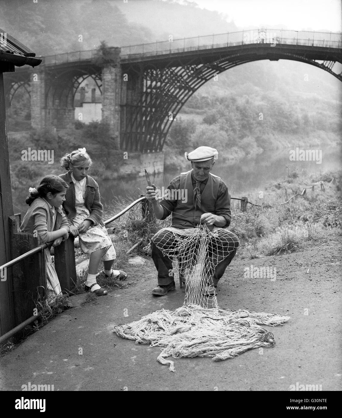 Coracle maker and poacher Harry Rogers repairing his fishing nets watched by two young girls in Ironbridge, Shropshire 1960 Britain Stock Photo