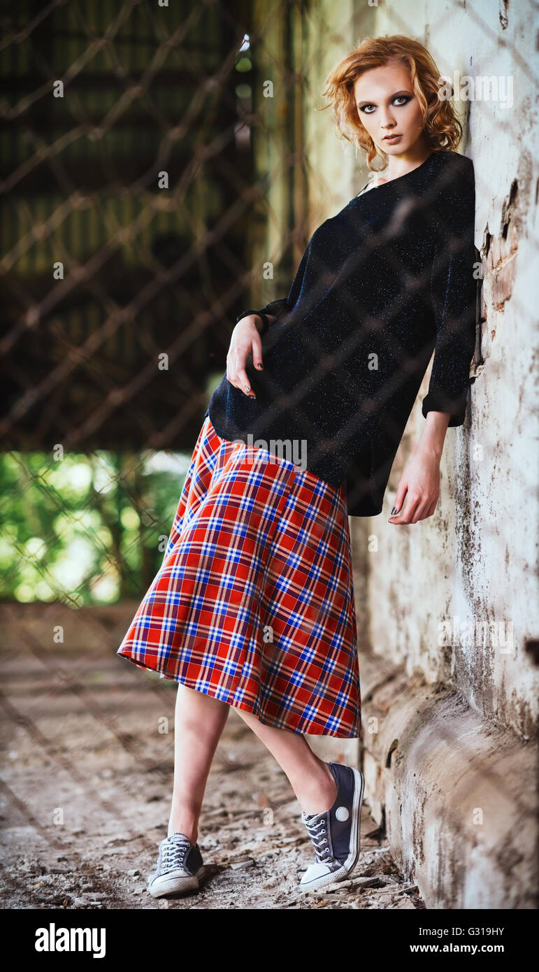 Grunge fashion: beautiful young girl in checkered skirt and blouse stands behind the lattice Stock Photo