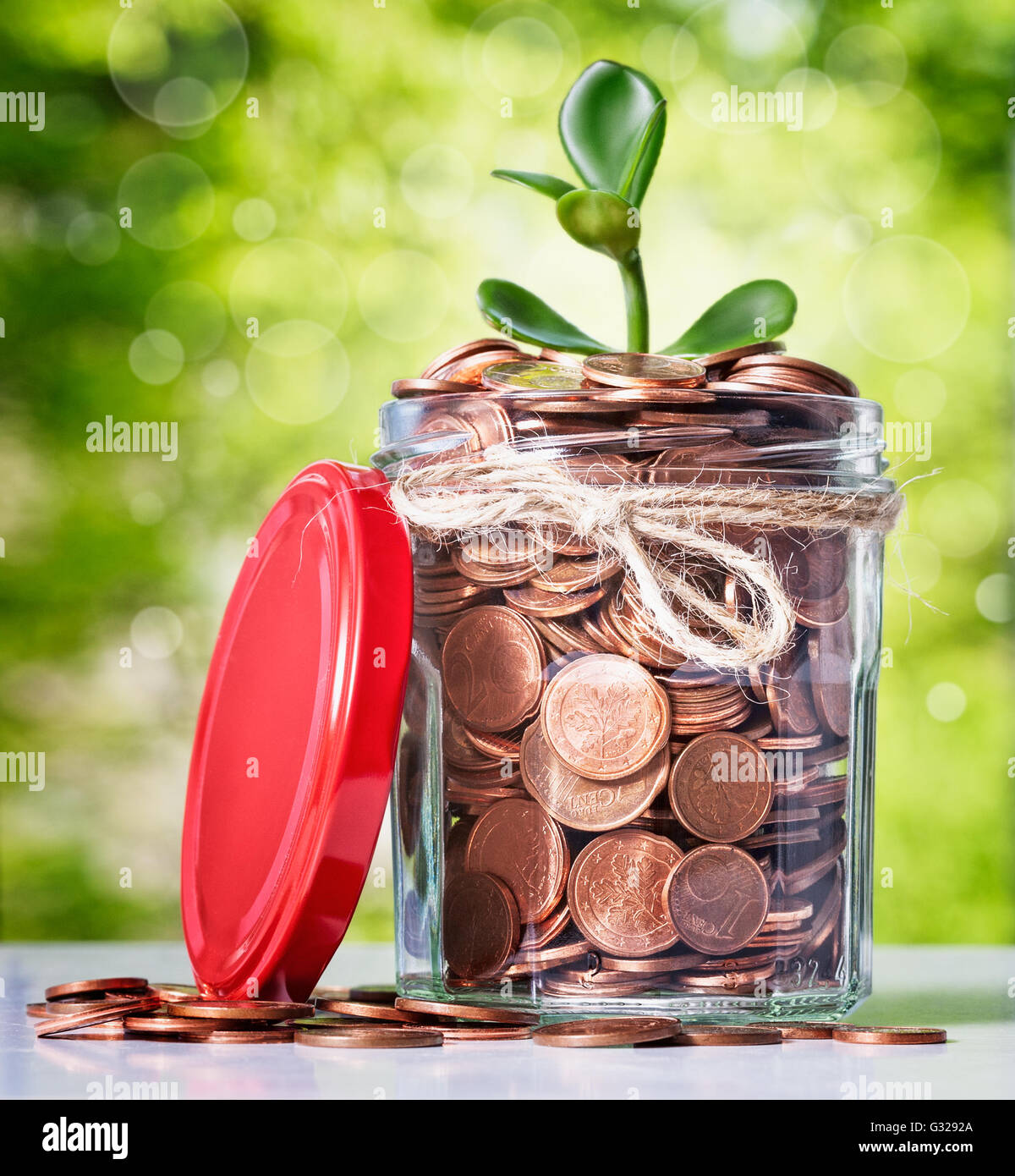 Glass bank jar with coins and growing sprout on green tree background. Saving money concept Stock Photo