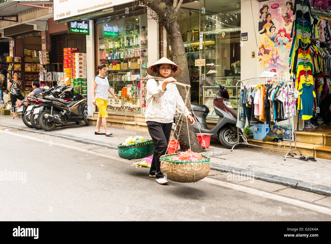 Street vendor carrying vegetables and fruit in baskets using a carrying pole, also called a shoulder pole in Hanoi Stock Photo