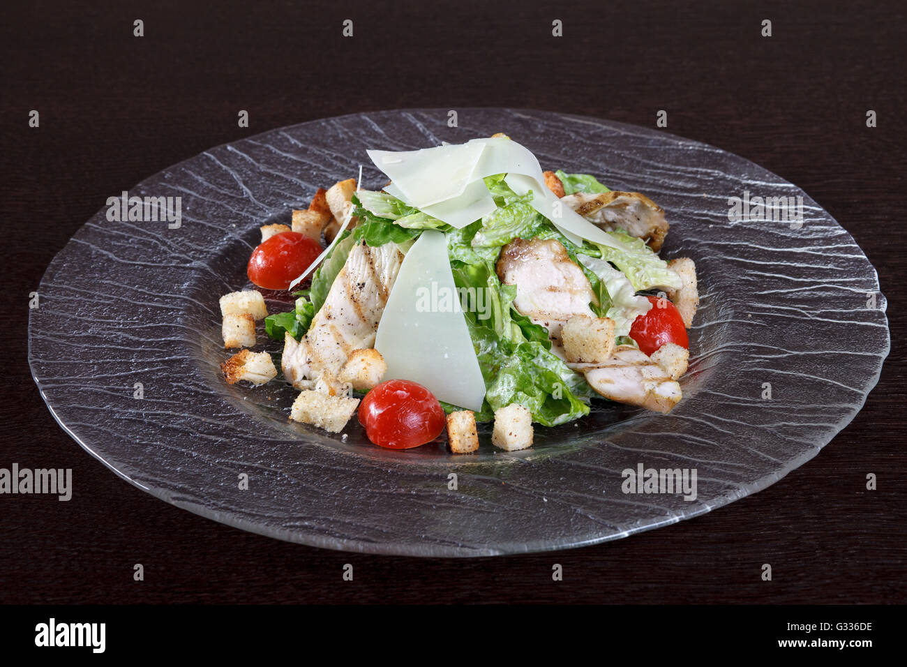 Salad with grilled chicken meat, lettuce, tomatoes and croutons. Dish of European cuisine. Glass plate on a brown wooden table.  Stock Photo