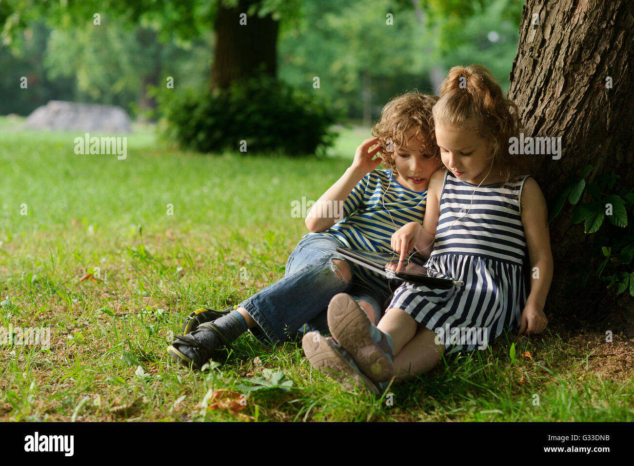 Boy and girl of 8-9 years sit on a grass in park with the small laptop. Children with interest look at the screen. Stock Photo