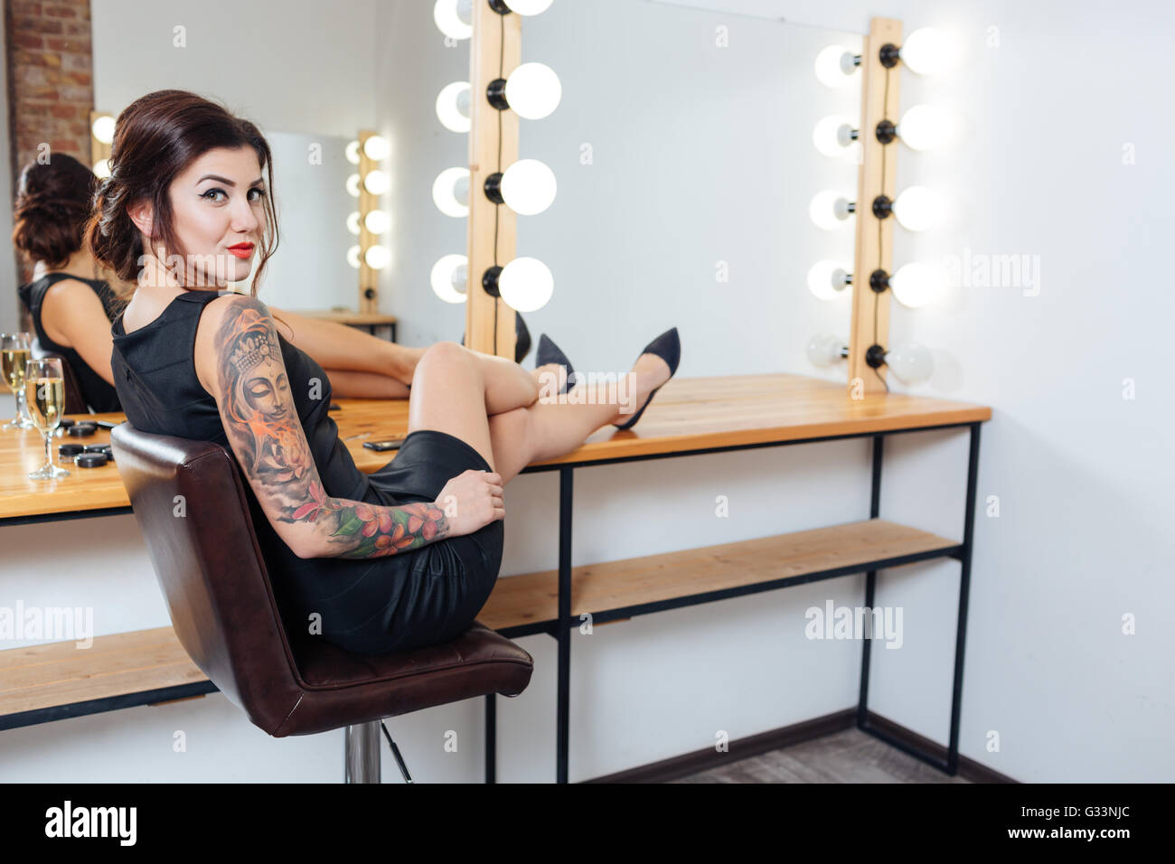 Happy tattooed young woman in black dress and shoes sitting in dressing room with hands on table Stock Photo