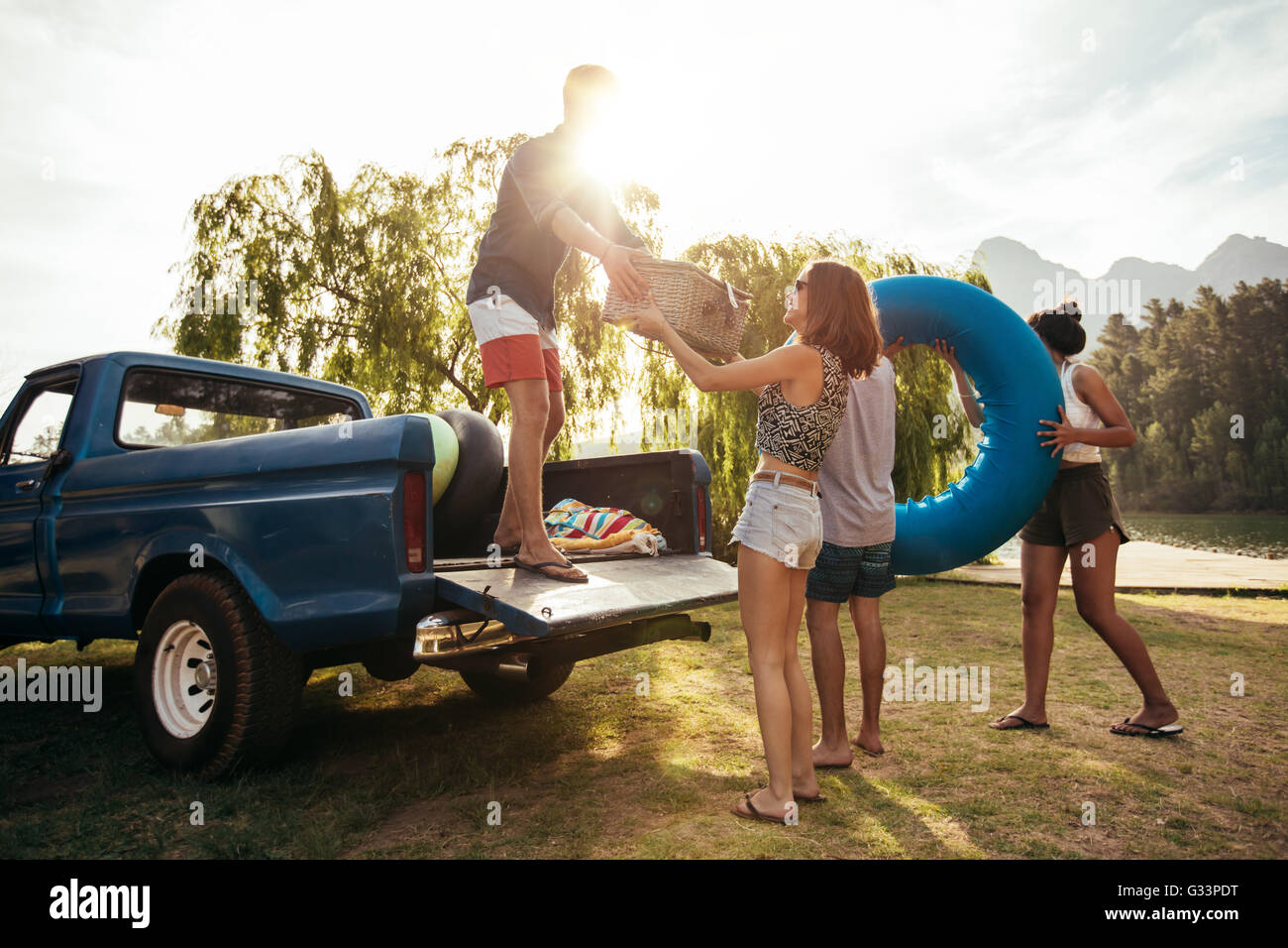Group of young friends on picnic by the lake. Men and women unloading pickup truck on camping trip, carrying picnic basket and i Stock Photo