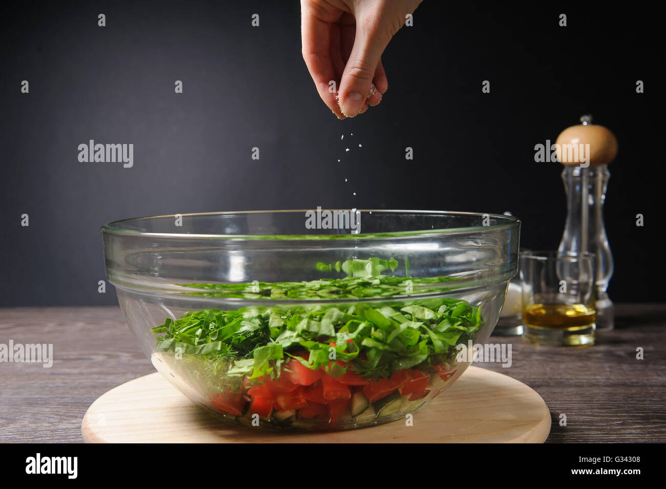 Hand adding salt to vegetable salad. Bowl of fresh green salad, tomatoes, cucumber on wood table against dark background. Stock Photo