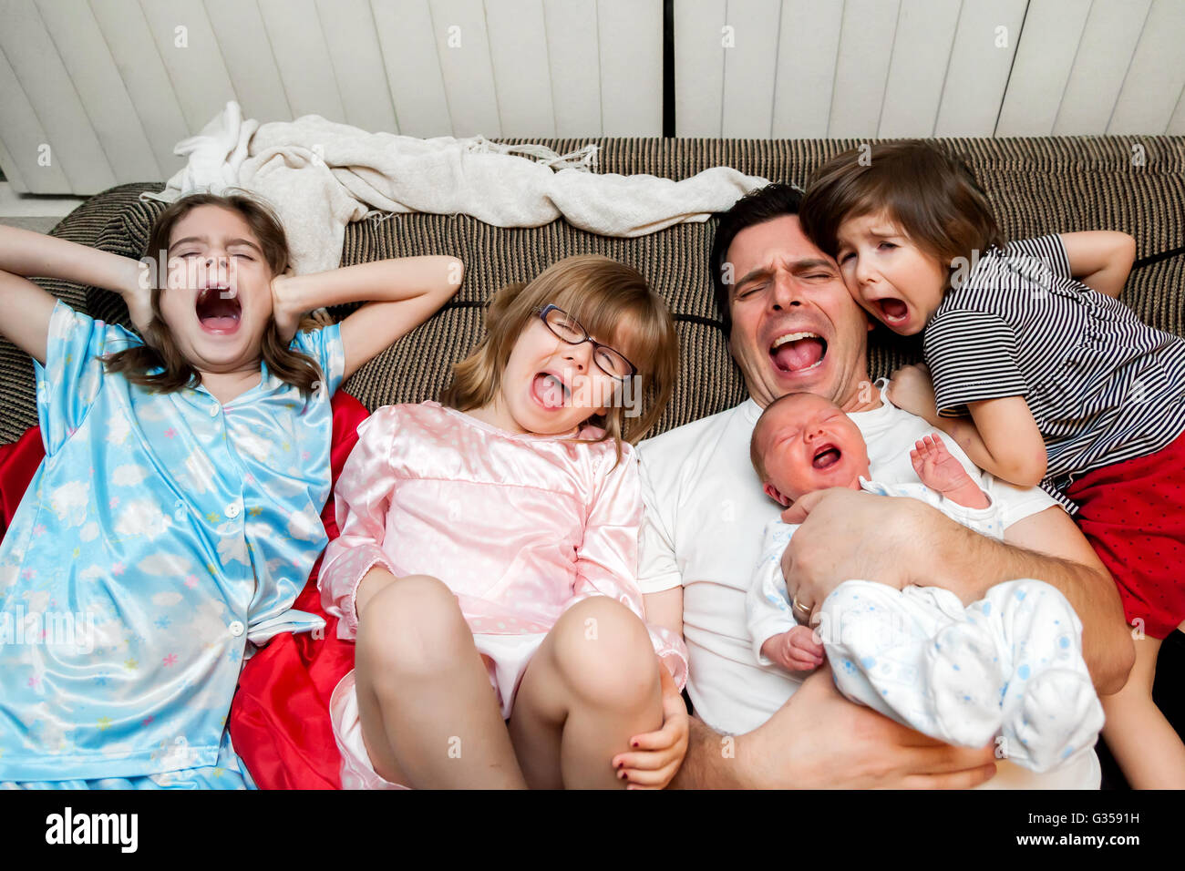 A father, his three daughters, and baby boy lean back on the couch with open mouth screams.  If you can’t beat them, join them. Stock Photo