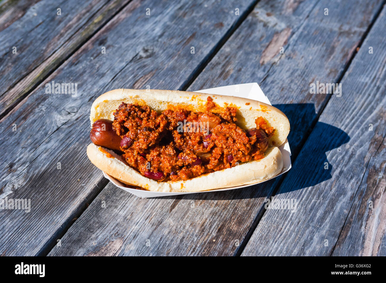 Sausage covered in hot chili on a bun in a paper tray, on weathered gray picnic table. Stock Photo