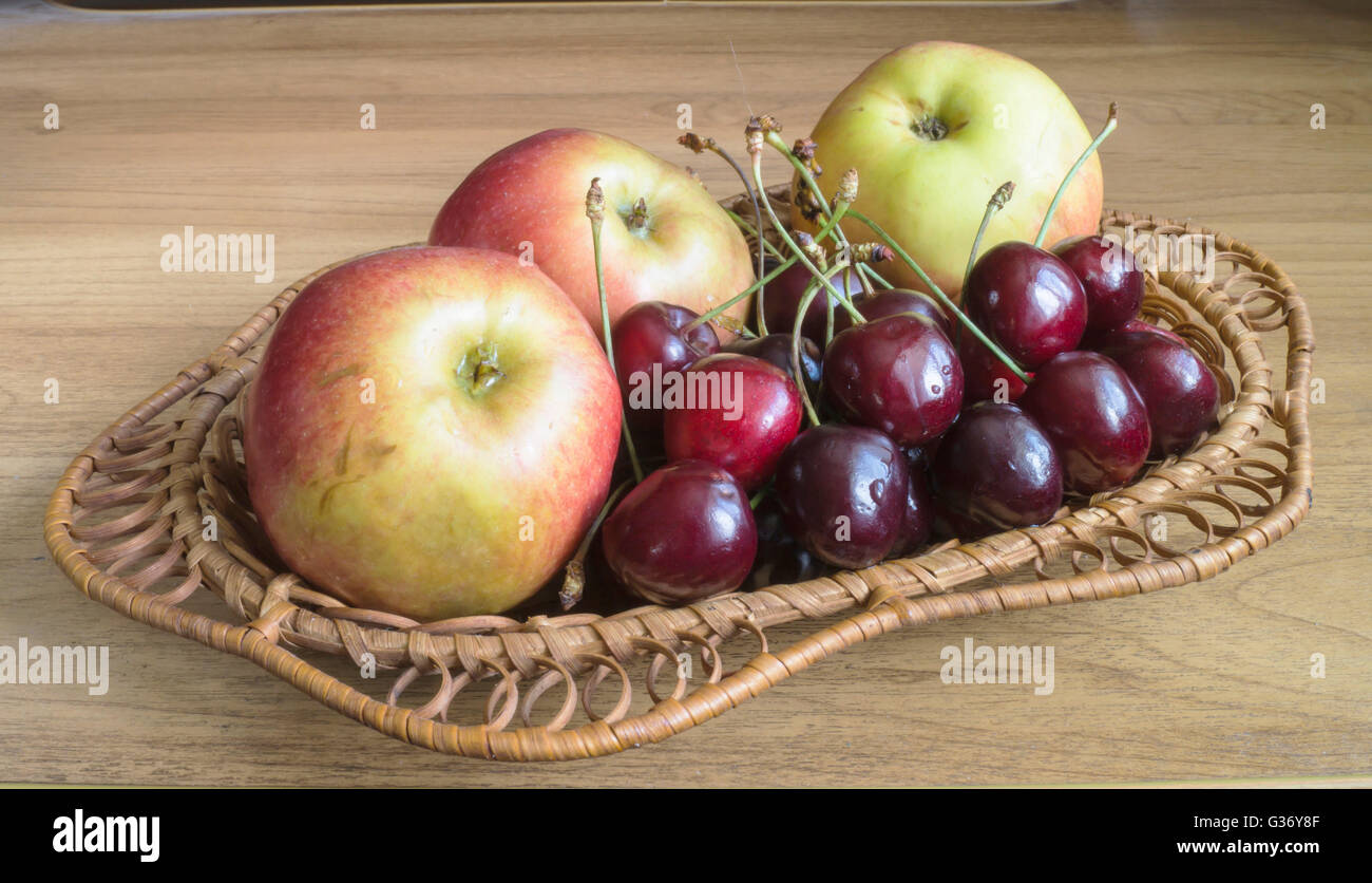 Ripe apple and sweet cherries in braided plate on wooden table Stock Photo