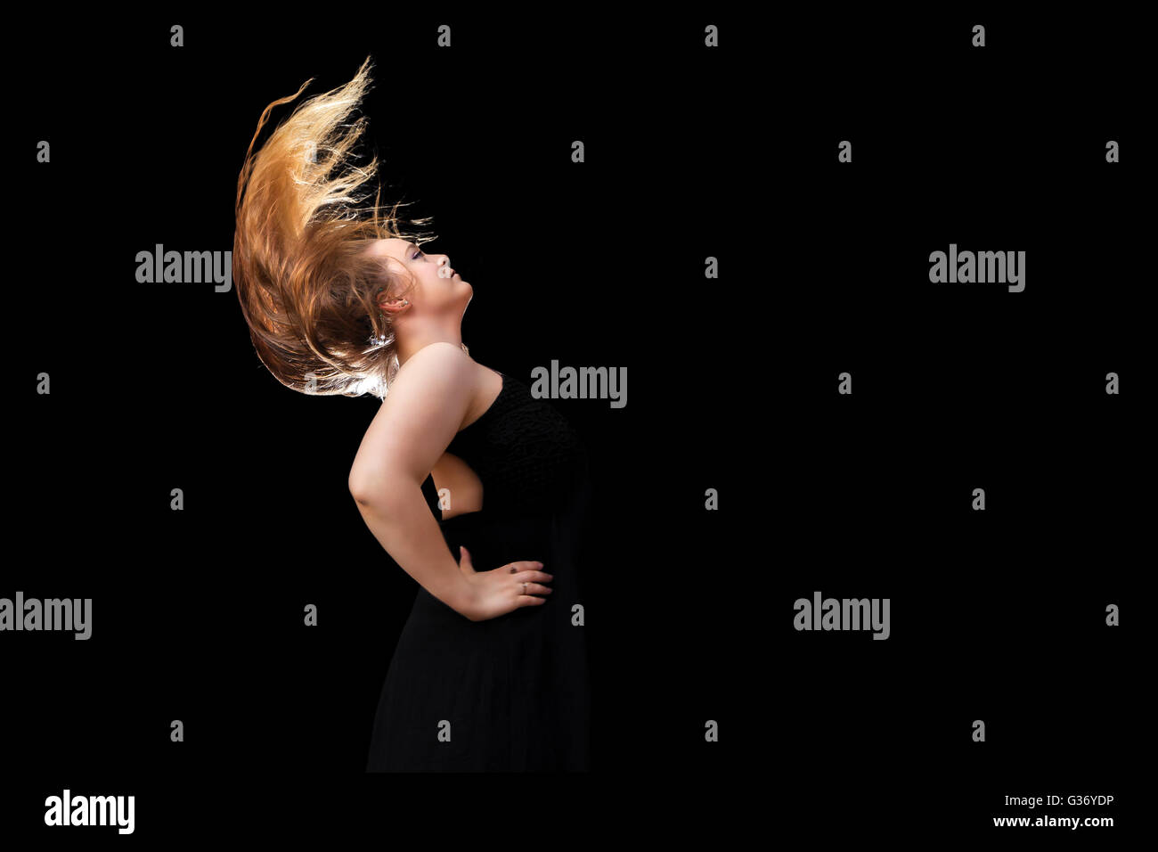 A teenage girl with beautiful, long, blond hair flips her hair against a black, background.  Her hair is back lit. Stock Photo