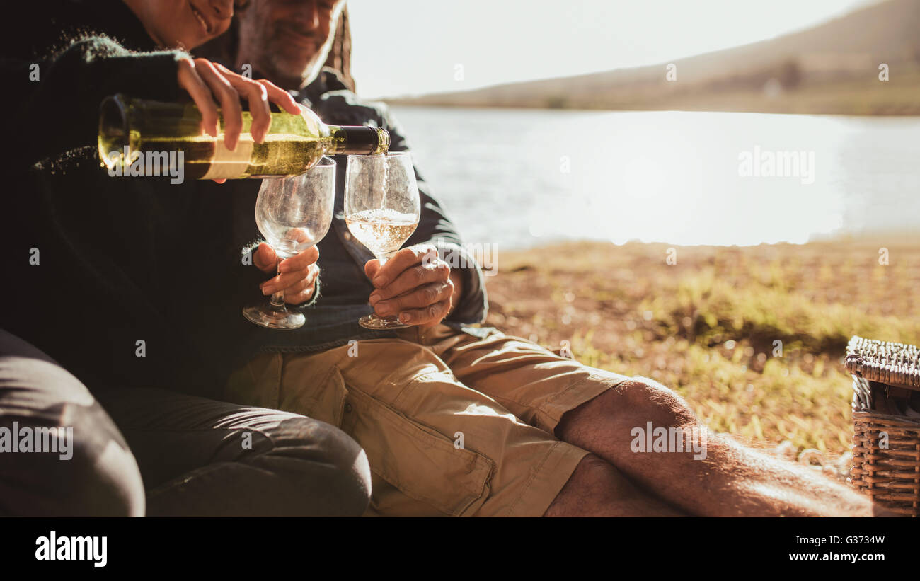 Cropped image of woman pouring wine in glasses. Couple camping near a lake enjoying with a glass of wine, focus on wine glasses. Stock Photo