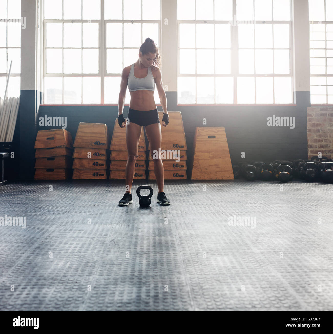 Full length shot of fit and strong woman standing in crossfit gym. Tough fitness female model with kettle bell on floor. Stock Photo