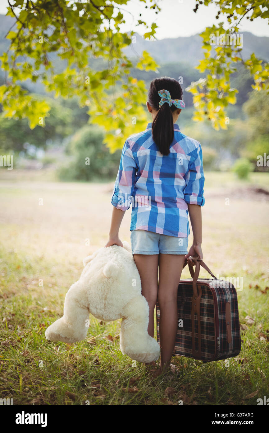 Girl standing in the park with a teddy bear and suitcases Stock Photo