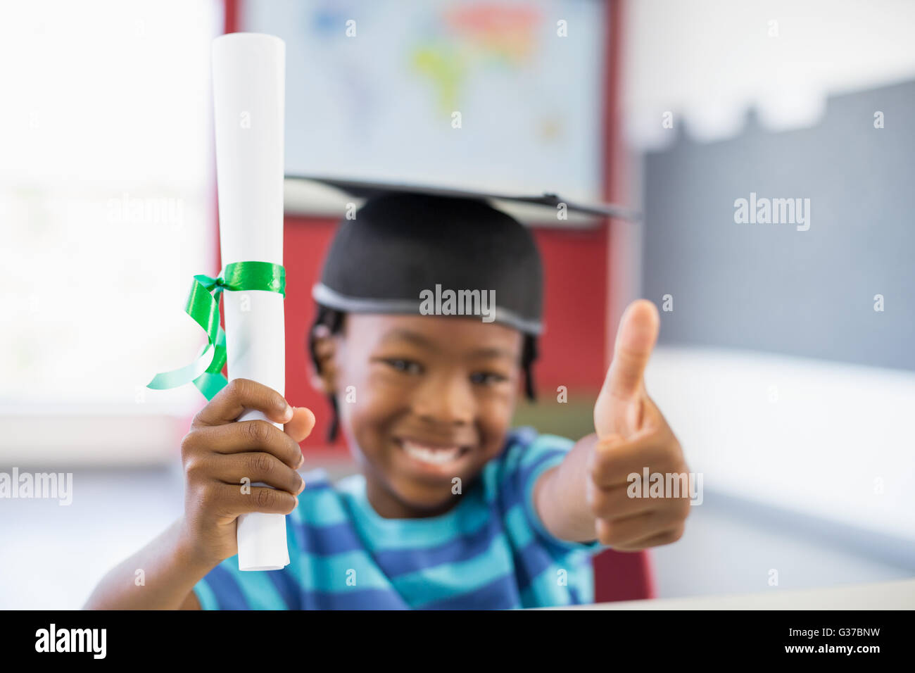Schoolboy in mortar board holding certificate and showing thumbs up in classroom Stock Photo