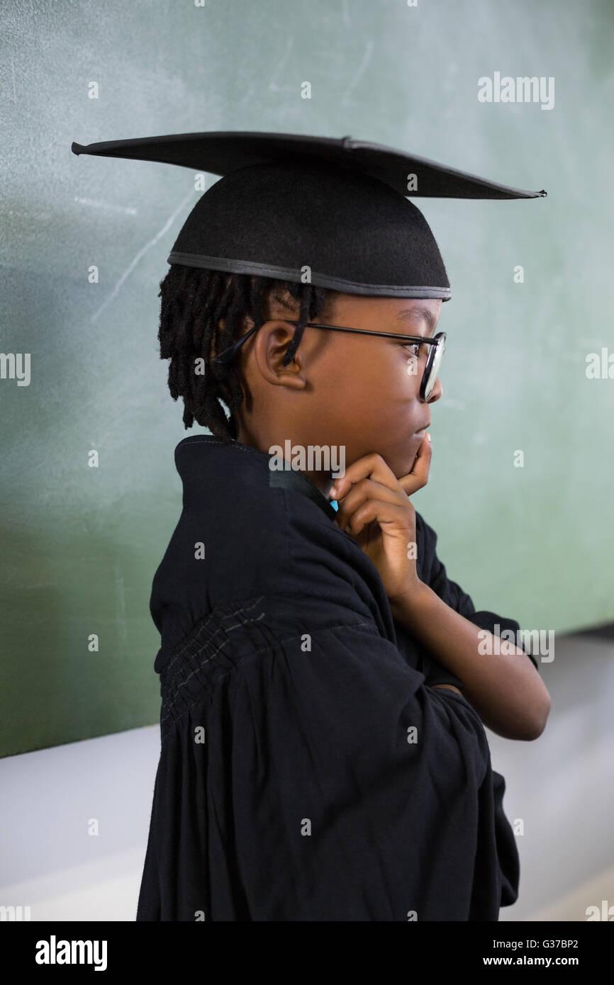 Thoughtful schoolboy wearing graduation gown in classroom Stock Photo