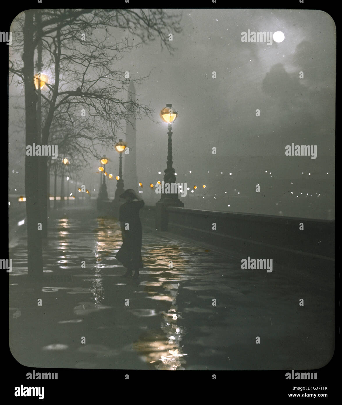 A woman walking along the  Embankment on a rainy night  with a full moon       Date: 1910 Stock Photo