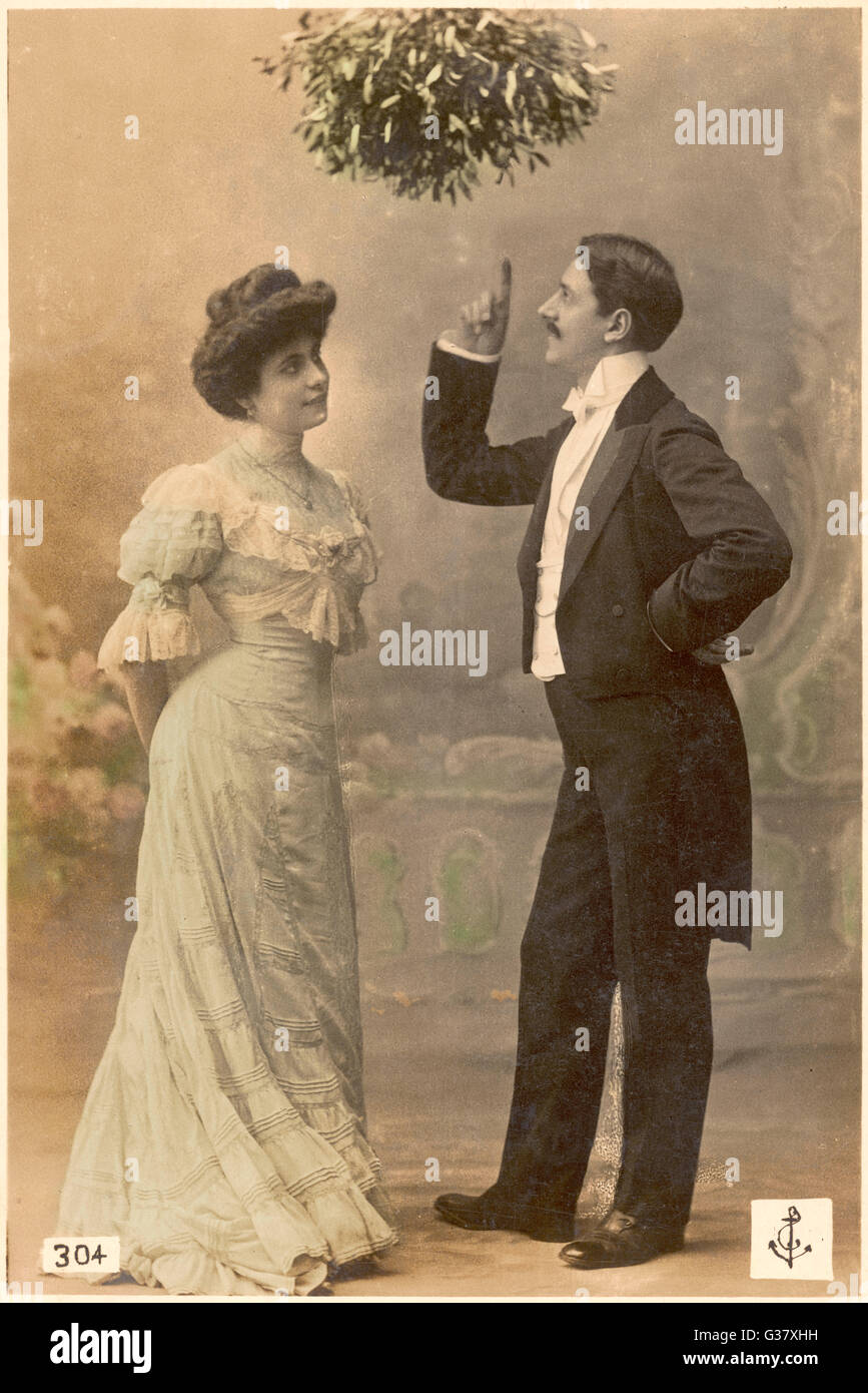 A gentleman treats a lady to a  brief lecture on the  significance of mistletoe ; she wonders when he will move  from theory to practice      Date: circa 1900 Stock Photo