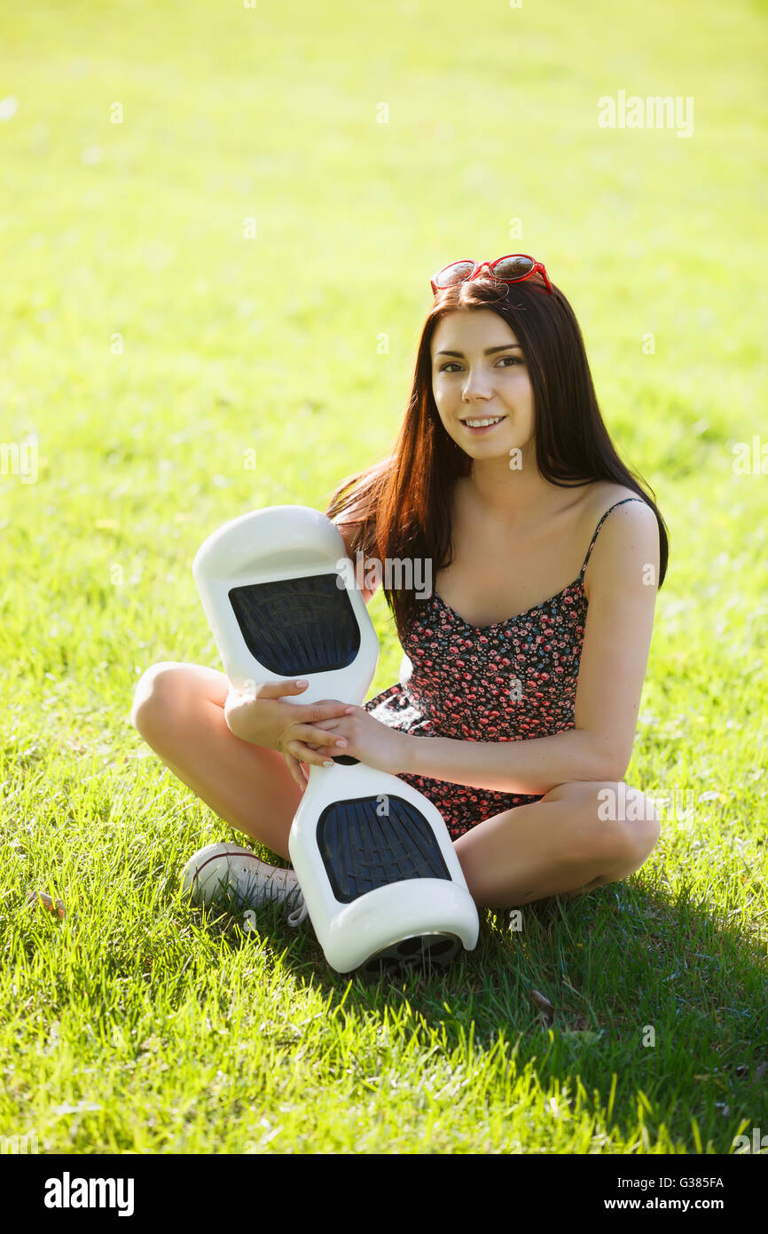 Young brunette girl with electrical mini hover board scooter in green park. Good summer weather, trendy transportation technology and cute model. Stock Photo