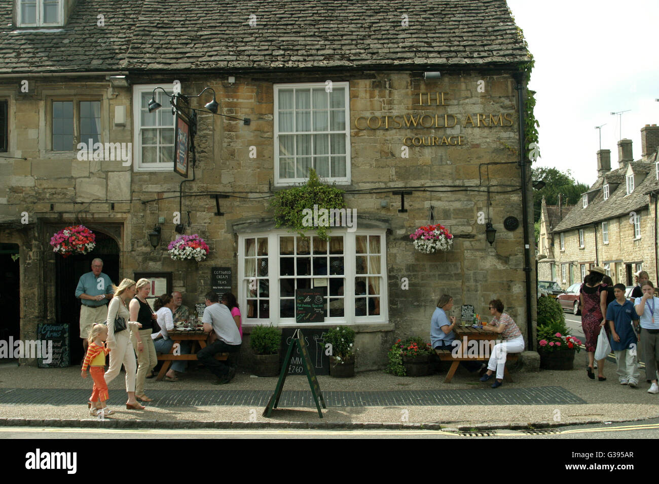 Cotswolds. The Cotswold Arms pub in full swing on 11th Century Burford's High Street with outdoor tables and passers-by. Stock Photo