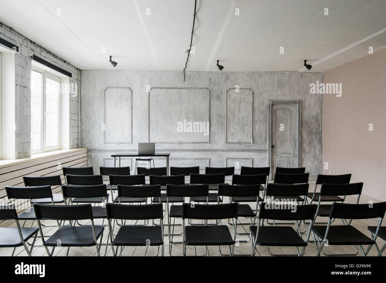 Room for lecture with a lot of dark chairs. Walls are white, loft interior. On the right there is a door. On the background there is a table with a laptop. Stock Photo