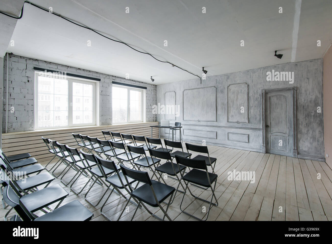 Room for lecture with a lot of dark chairs. Walls are white, loft interior. On the right there is a door. On the background there is a table with a laptop. Stock Photo