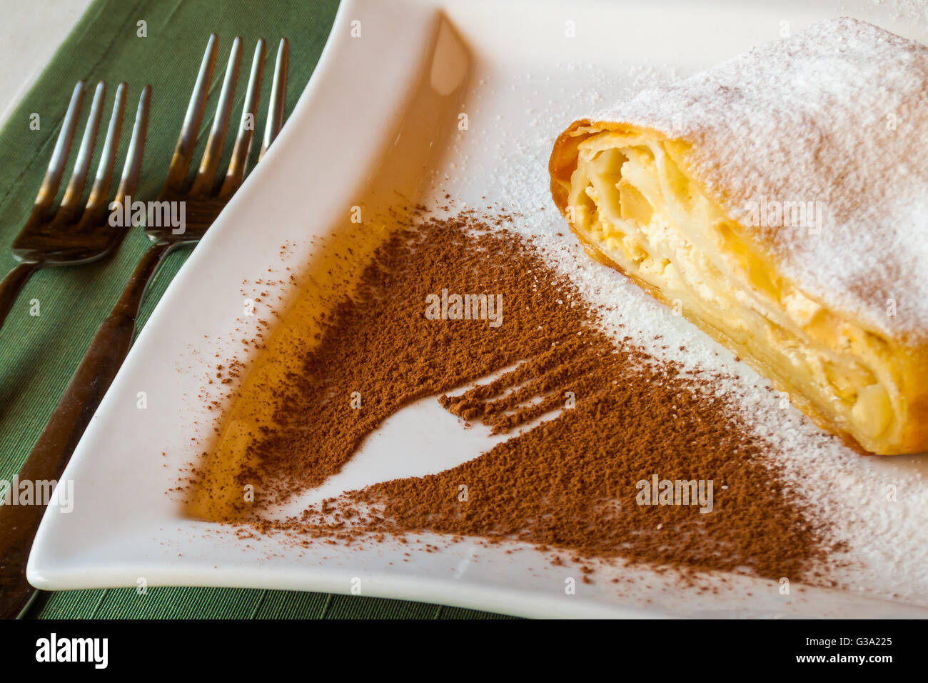 delicious strudel stuffed with cottage cheese, assign with powdered sugar on a white plate with a fork pattern of cocoa powder. Stock Photo