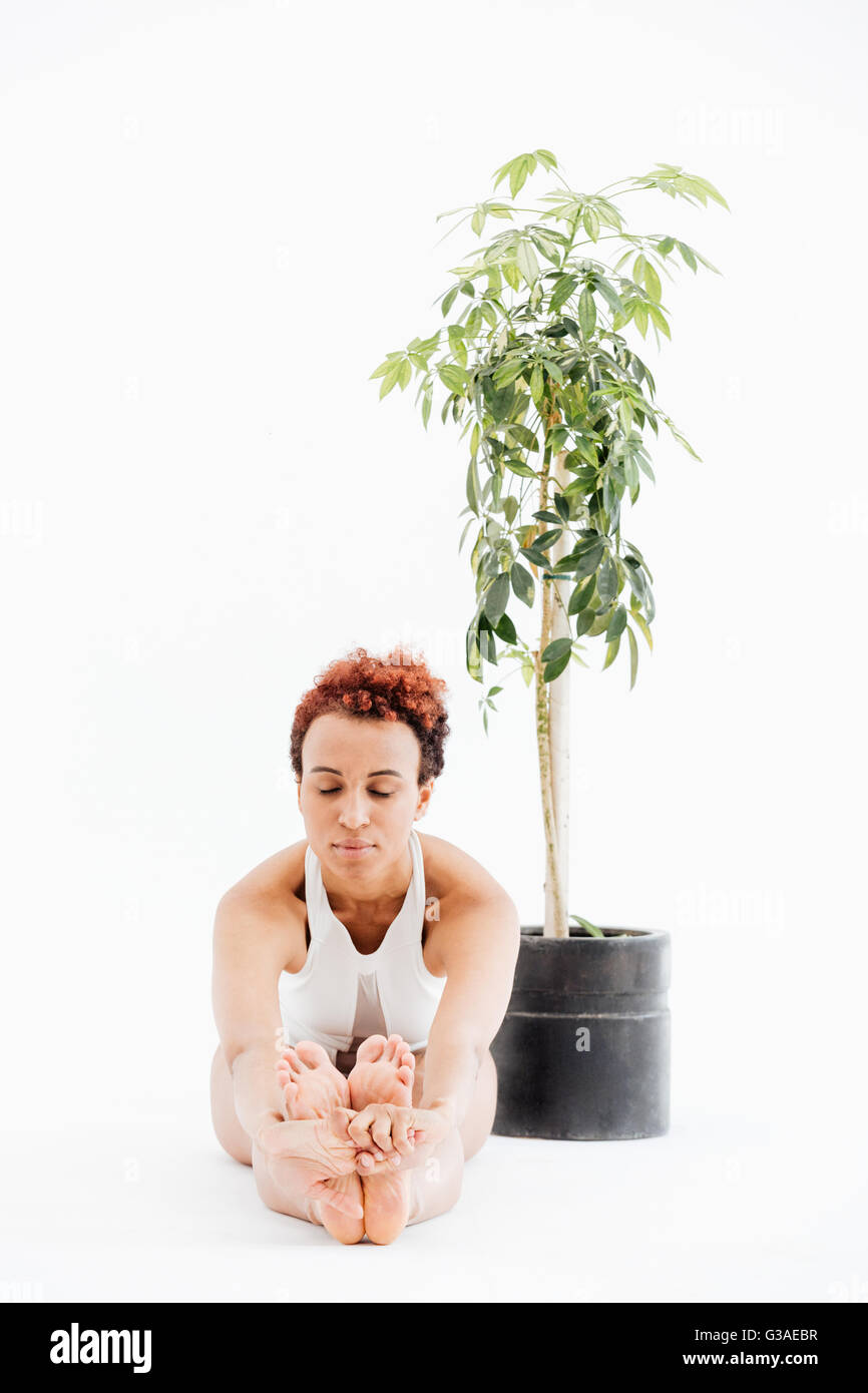Peaceful african american young woman sitting near the tree in pot and doing yoga exercises over white background Stock Photo